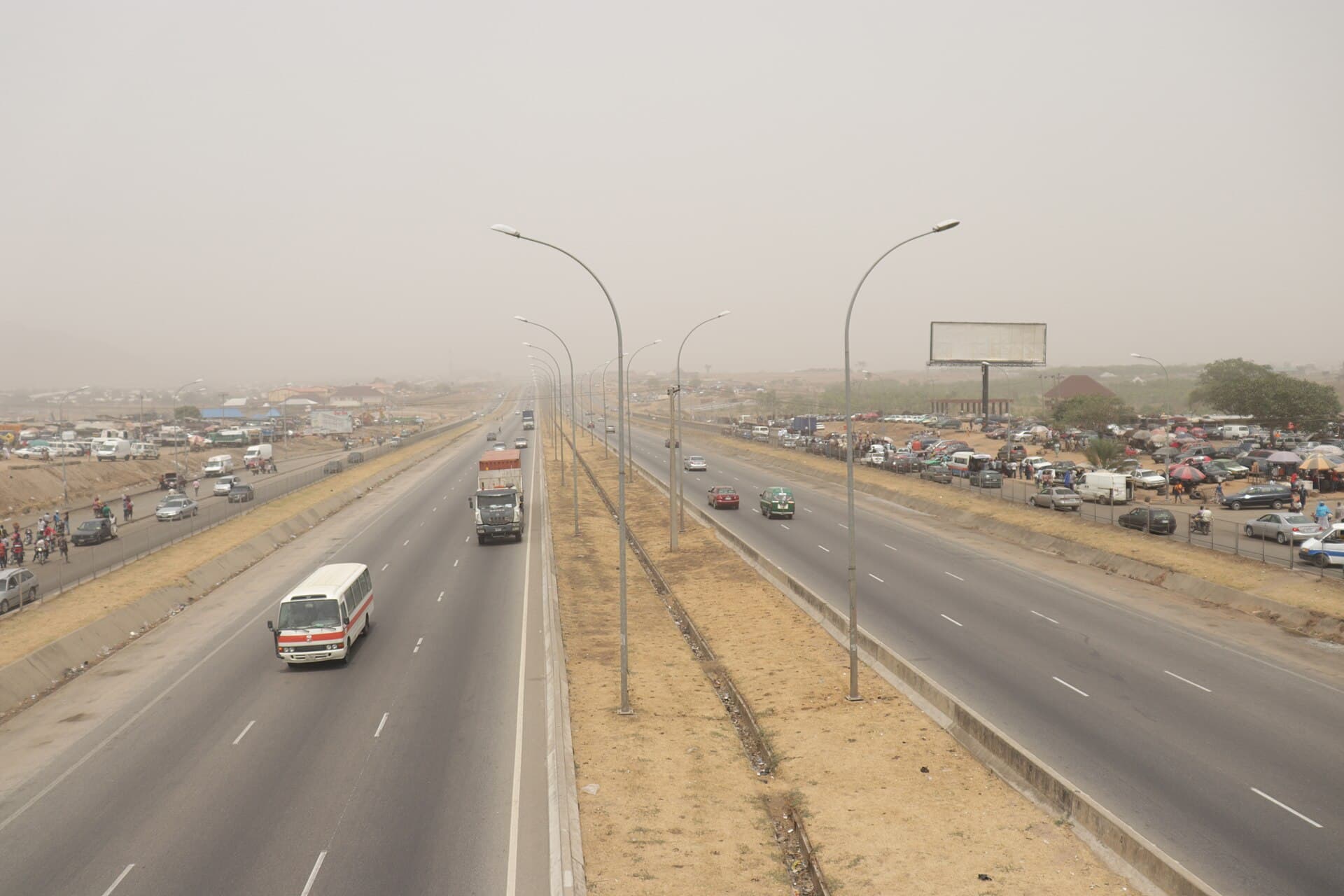 A Nigerian expressway flattened by harmattan haze, traffic thinning toward the horizon