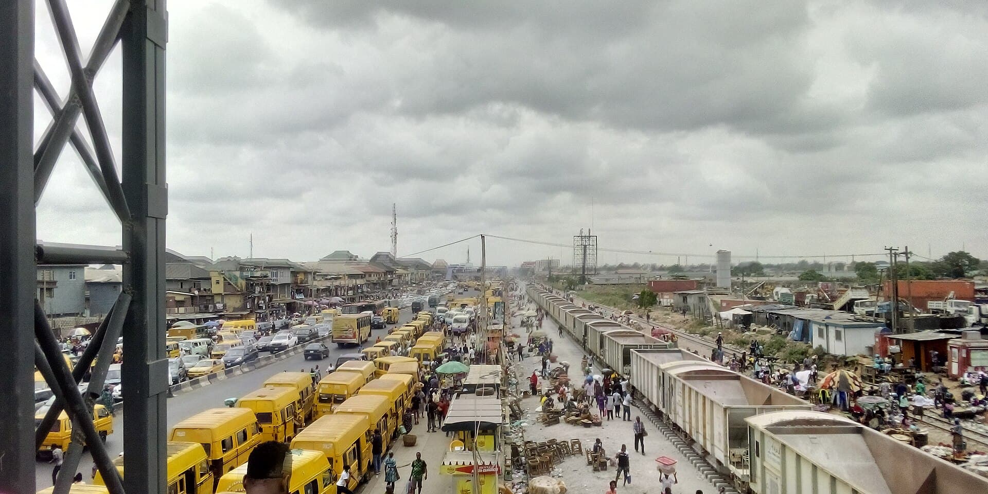 Danfo buses banked up along a Lagos expressway beside a freight line, market stalls spilling onto the tracks
